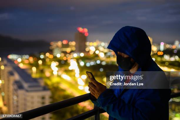 man in hooded sweater holding a mobile phone in dark with city in background - blanqueo de dinero fotografías e imágenes de stock