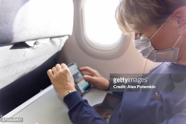 international flight.  mature women in face mask looking photos on her smartphone in airplane. - alex potemkin coronavirus stock pictures, royalty-free photos & images