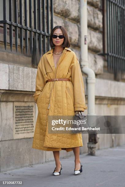 Aimee Song wears sunglasses, a yellow long padded trench coat, a brown leather Hermes belt, a dark brown Hermes bag, brown and white pointed shoes,...