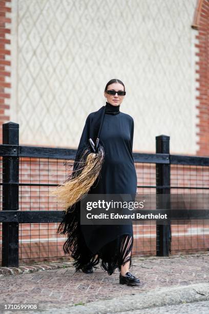 Guest wears sunglasses, a black hi-neck wide fringy dress, a matching large shawl, black moccasins, outside Alberta Ferretti, during the Milan...