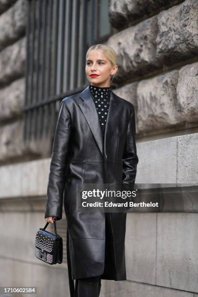 Caroline Daur wears sunglasses, a black long leather trench coat, a black turtleneck studded dress, knee-high leather pointed boots, outside Hermes,...