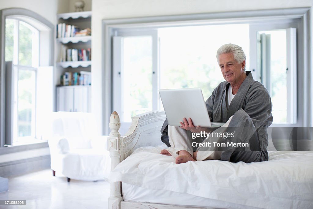 Uomo anziano in accappatoi usando il portatile in camera da letto