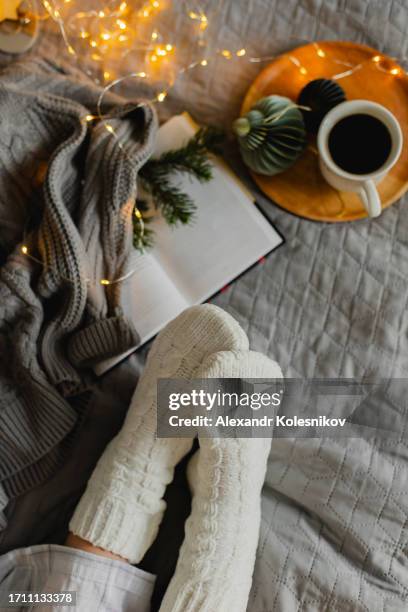 top view of female feet in cozy white socks on bed with cup of coffee, christmas lights and open book. christmas, relaxation and hygge concept. - cozy winter stock pictures, royalty-free photos & images