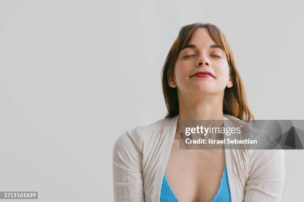 young beautiful woman with closed eyes inhaling fresh air relaxing deeply in front of white cloth background. - zen-like stock pictures, royalty-free photos & images