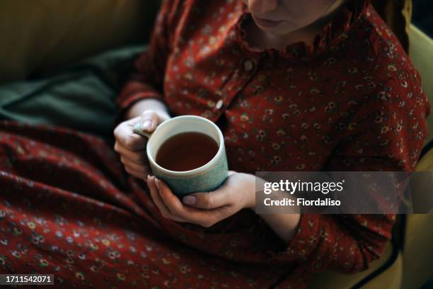 portrait of a long-haired red-haired woman sitting on the sofa with cup of tea - krankenstand stock-fotos und bilder