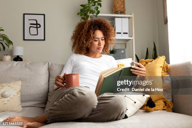 young multiracial woman reading a book at home, drinking tea sitting on the sofa in cozy living room. - lit photos et images de collection