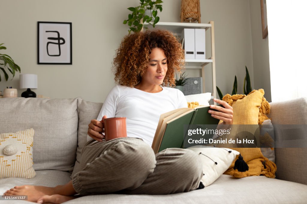 Young multiracial woman reading a book at home, drinking tea sitting on the sofa in cozy living room.