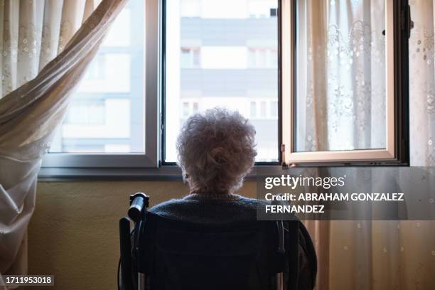 una donna anziana seduta su sedia a rotelle che guarda fuori dalla finestra di casa - longevità foto e immagini stock