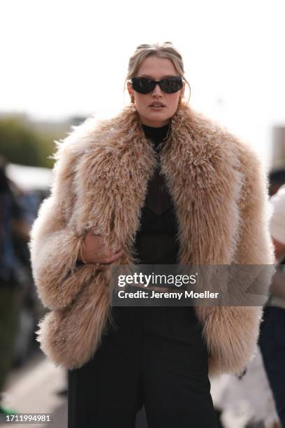 Toni Garrn is seen wearing a brown oversized fur jacket, with a black transparent top, black bralette, high-cut black pants and black sunglasses...