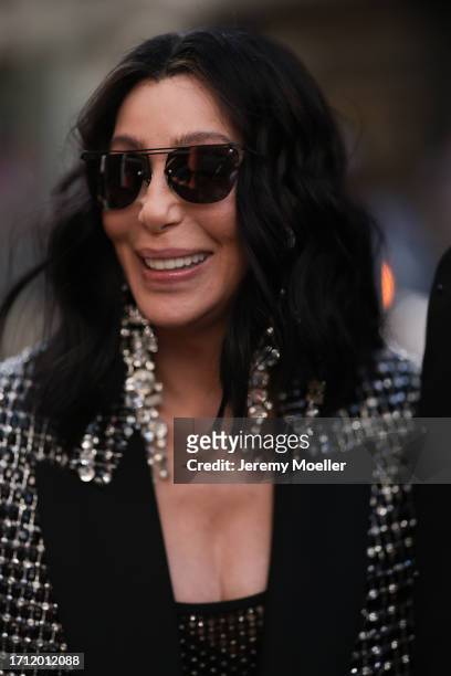 Cher is seen wearing dark shades and big silver diamond earrings, a black top with silver ornaments and matching oversized black jacket with silver...