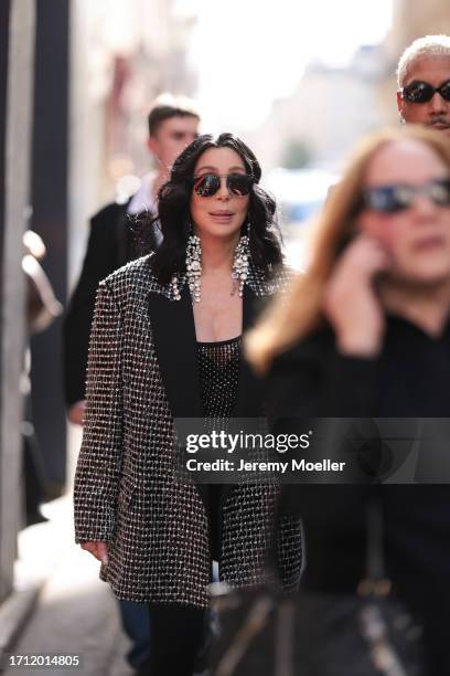 Cher is seen wearing dark shades and big silver diamond earrings, a black top with silver ornaments and matching oversized black jacket with silver...