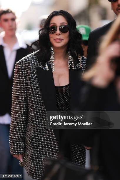 Cher is seen wearing dark shades and big silver diamond earrings, a black top with silver ornaments and matching oversized black jacket with silver...