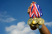 Athlete Hand Holding Up Bunch of Gold Medals Blue Sky