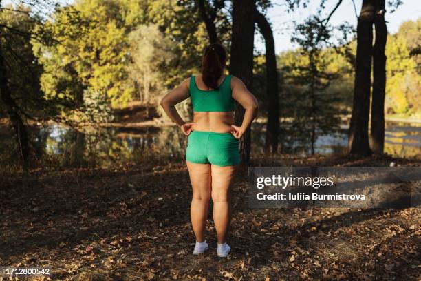 confident woman in green sports attire exposing plump body showcases love handles facing serene river - man standing full body stock-fotos und bilder