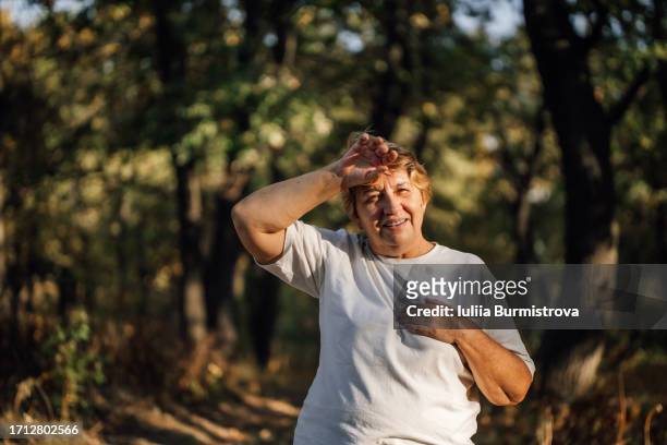 cheerful senior lady in white t-shirt stands among trees feeling tired and out of breath - overheated stock pictures, royalty-free photos & images