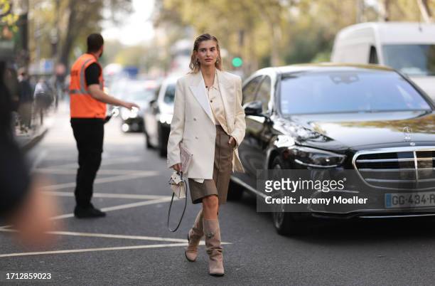 Xenia Adonts is seen outside Hermes show wearing silver earrings, beige silk blouse, white oversized blazer, long shorts in brown with matching...