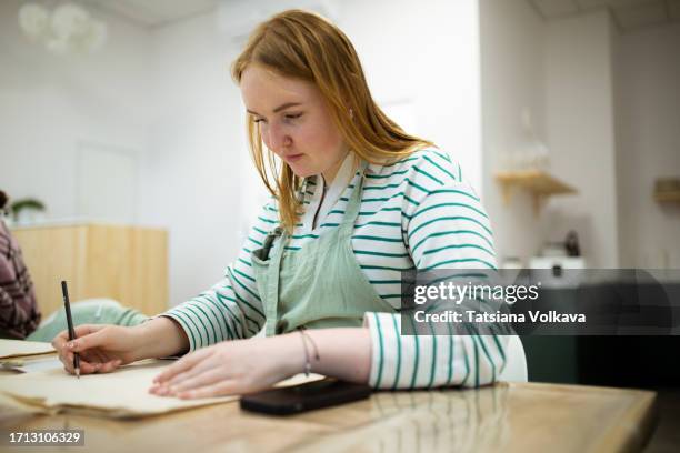 beautiful young woman with blonde hair delicately sketches artistic vision onto beige shopping bag - brightly lit classroom stock pictures, royalty-free photos & images