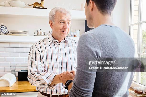 senior father sharing good news with adult son in kitchen - secret handshake stock pictures, royalty-free photos & images