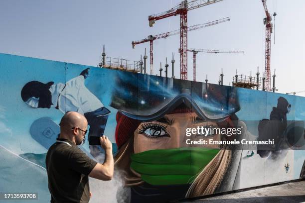 Man uses his mobile phone to take a picture of a mural representing snowboarders and painted on the outer wall of the building site of the 2026...