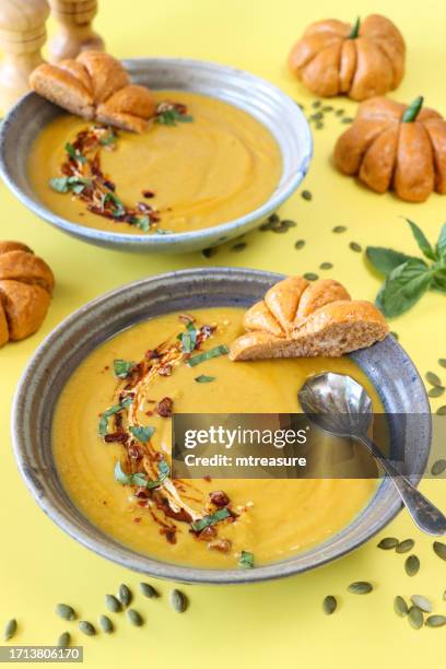 close-up image of bowls filled with homemade halloween pumpkin soup, yoghurt and red chilli garnish, pumpkin seeds, metal spoon, pumpkin designed bread rolls with green chilli pepper stalk, yellow background, elevated view, focus on foreground - soup bowl stock pictures, royalty-free photos & images