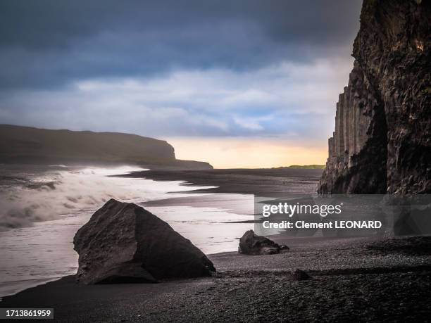 in-depth view of the reynisfjara black sand beach at sunset during a cloudy day, with the basalt columns and basalt cave of the reynisfjall mountain on the right side, vík í mýrdal (vik), south iceland (suðurland). - boulders beach stock pictures, royalty-free photos & images