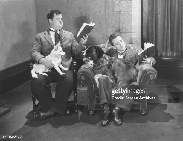 Stan Laurel and Oliver Hardy relax in armchairs with dogs on their laps as they read books in a portrait of the comedy duo circa 1930.