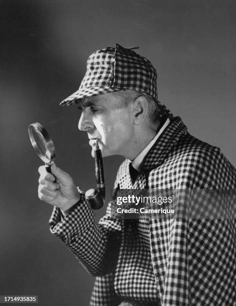 Older man in a Sherlock Holmes type matching outfit and hat with a hand and a magnifying glass up to his face with a large pipe hanging out of his...