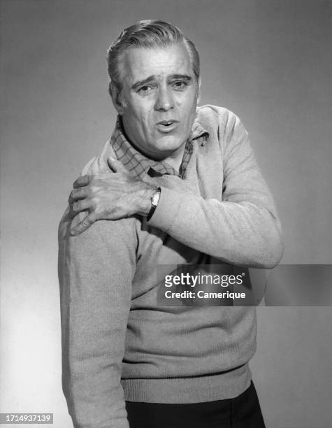 Older man in a pullover sweater posed looking at the camera with a facial expression of pain while holding one arm down and the other one over...