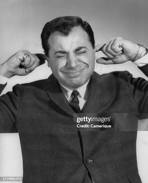Middle-aged businessman in a suit and tie with his fingers in his ears and a grimacing look on his face as he attempts to block something out.