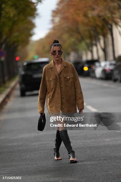 Alexandra Pereira wears sunglasses, a brown suede oversized jacket, a white pleated mini skirt, a leather bag, knee-high black leather boots with...