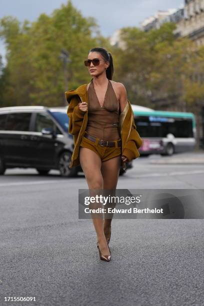 Camila Coelho wears sunglasses, a brown ribbed corduroy jacket, a v-neck low neck mesh sleeveless body suit, a leather brown belt, mini shorts,...