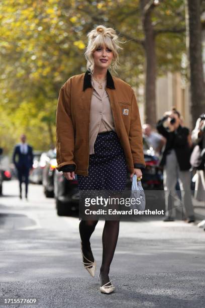 Emili Sindlev wears a brown Carhartt jacket, a beige cardigan, a mini skirt in dark blue with printed polka dots, black tights, a pastel pale bag...