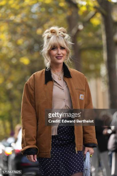 Emili Sindlev wears a brown Carhartt jacket, a beige cardigan, a mini skirt in dark blue with printed polka dots, black tights, a pastel pale bag...