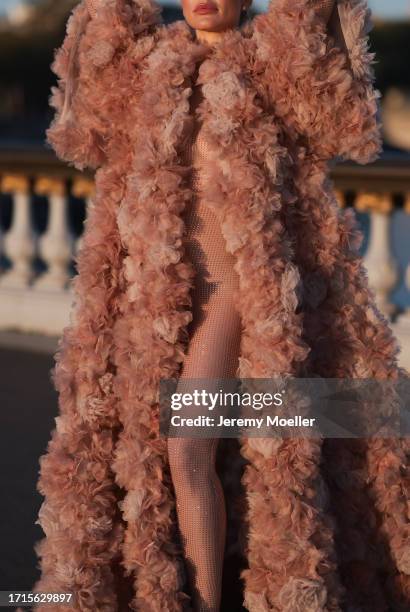 Ginta is seen wearing a voluminous coat with train, covered with beige and rose tulle flowers all over and underneath a tight, beige, transparent,...