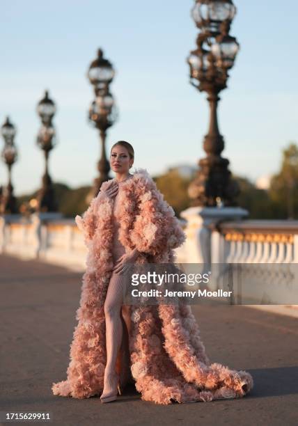 Ginta is seen wearing a voluminous coat with train, covered with beige and rose tulle flowers all over and underneath a tight, beige, transparent,...