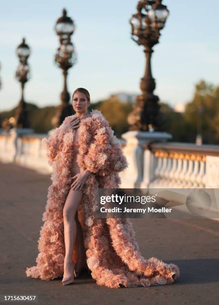 Ginta is seen wearing a voluminous coat with train, covered with beige and rose tulle flowers all over and underneath a tight, beige, transparent,...