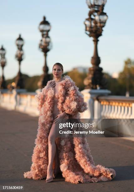 Ginta is seen wearing a voluminous coat with train, covered with beige and rose tulle flowers all over and underneath a tight, beige, transparent,...