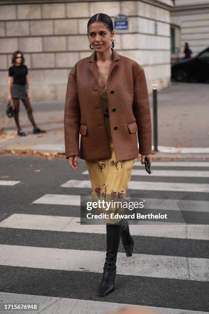 Guest wears earrings, a brown leather texture jacket, a floral print flowing dress, black leathe rboots, outside Miu Miu, during the Womenswear...