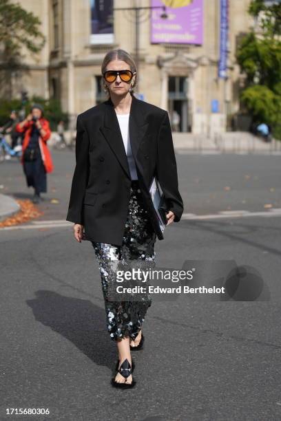 Guest wears orange sunglasses, a white t-shirt, a black oversized blazer jacket, a midi silver shiny sequined skirt, sandals, outside Miu Miu, during...