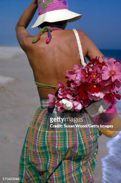 An unidentified female model walks along the beach, dressed in a crochet hat and swimsuit, and multi-colored Tartan skirt, with a crochet bag full of...