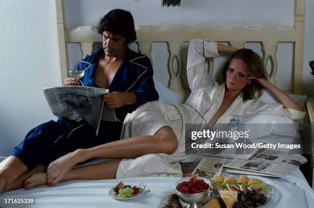 Portrait of American model and actress Cheryl Tiegs and her husband, film director Stan Dragoti, lie in bed with newspapers and food from room...