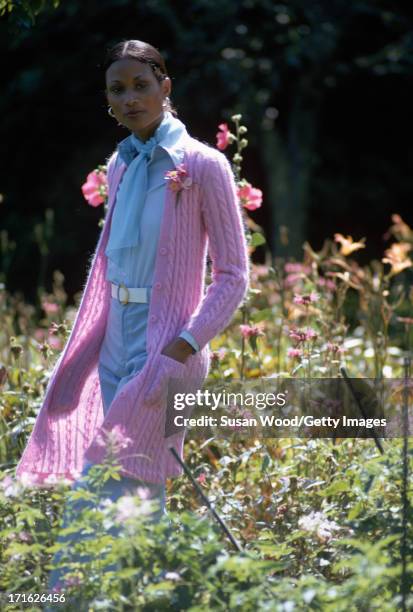 Portrait of an unidentified female model as she poses amid the flowers of a garden, dressed in a full-length, pink cable-knit cardigan and matching,...