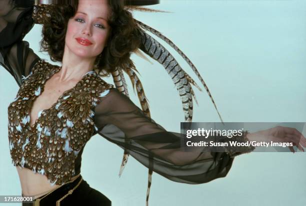 Portrait of an unidentified female model, dressed in a sheer blouse decorated with applique feathers, as she poses against a white background with a...