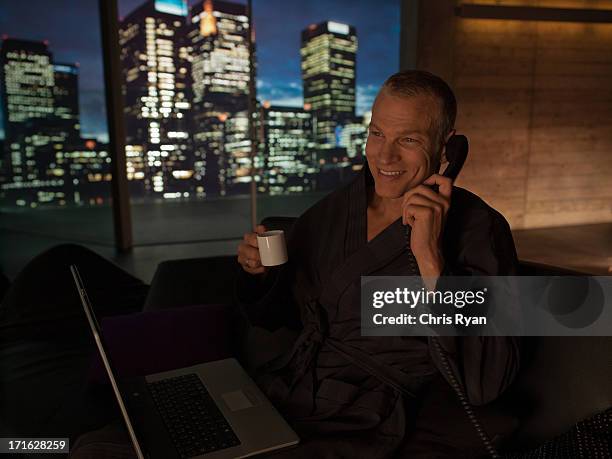 man in bathrobe with laptop drinking coffee and talking on telephone - landline phone home stock pictures, royalty-free photos & images