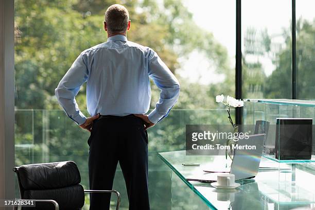 businessman looking out office window - handen op de heupen stockfoto's en -beelden