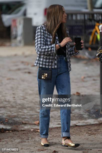 Guest wears a black and white checked tweed jacket, a black quilted leather bag, blue hems denim jeans, golden flat pointed shoes, outside Chanel,...