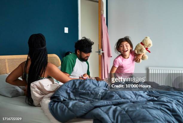 young girl smiling and holding teddy bear, parents in bed - waking up stock pictures, royalty-free photos & images
