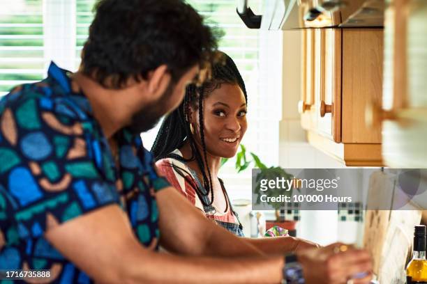 black woman smiling towards man in kitchen as they prepare food - woman 30s house busy stock pictures, royalty-free photos & images