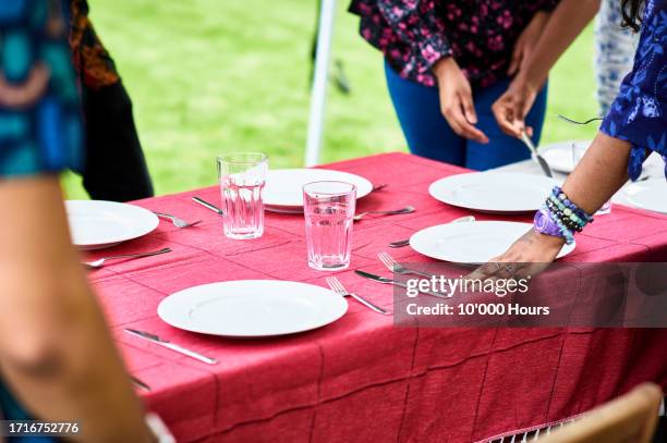 people setting out plates and cutlery on table with red tablecloth outdoors - place setting stock pictures, royalty-free photos & images