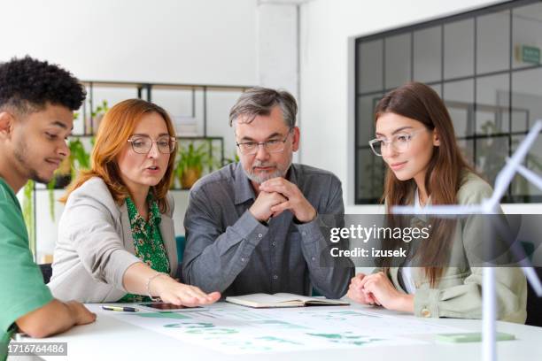 business colleagues discussing over graph while sitting at desk with wind turbine models in office - sustainable business stock pictures, royalty-free photos & images